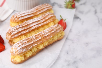 Delicious eclairs filled with cream and strawberries on white marble table, space for text