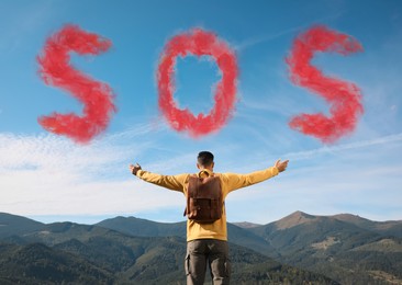 Man with backpack and word SOS made of color smoke bomb in mountains, back view