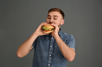 Young man eating tasty burger on grey background