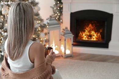 Photo of Woman with hot drink resting near fireplace in cozy room decorated for Christmas, back view