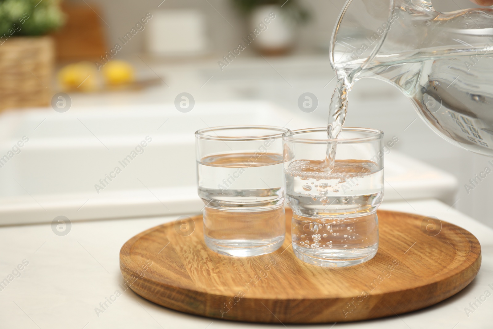 Photo of Pouring water from jug into glass on white table in kitchen, closeup. Space for text