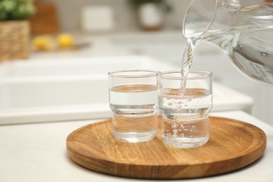 Pouring water from jug into glass on white table in kitchen, closeup. Space for text