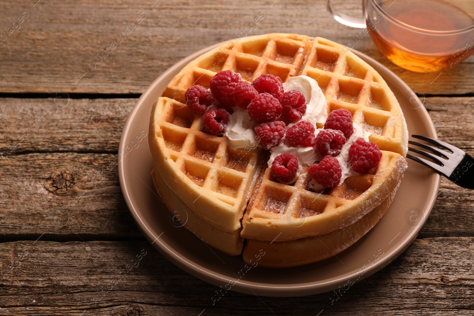 Photo of Tasty Belgian waffles with fresh raspberries, whipped cream, cup of tea and fork on wooden table, closeup