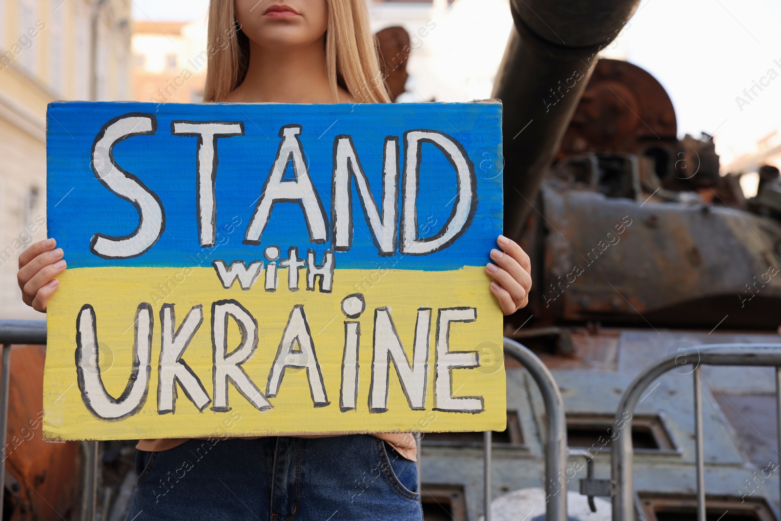 Photo of Woman holding poster in colors of national flag with words Stand with Ukraine near broken tank on city street, closeup