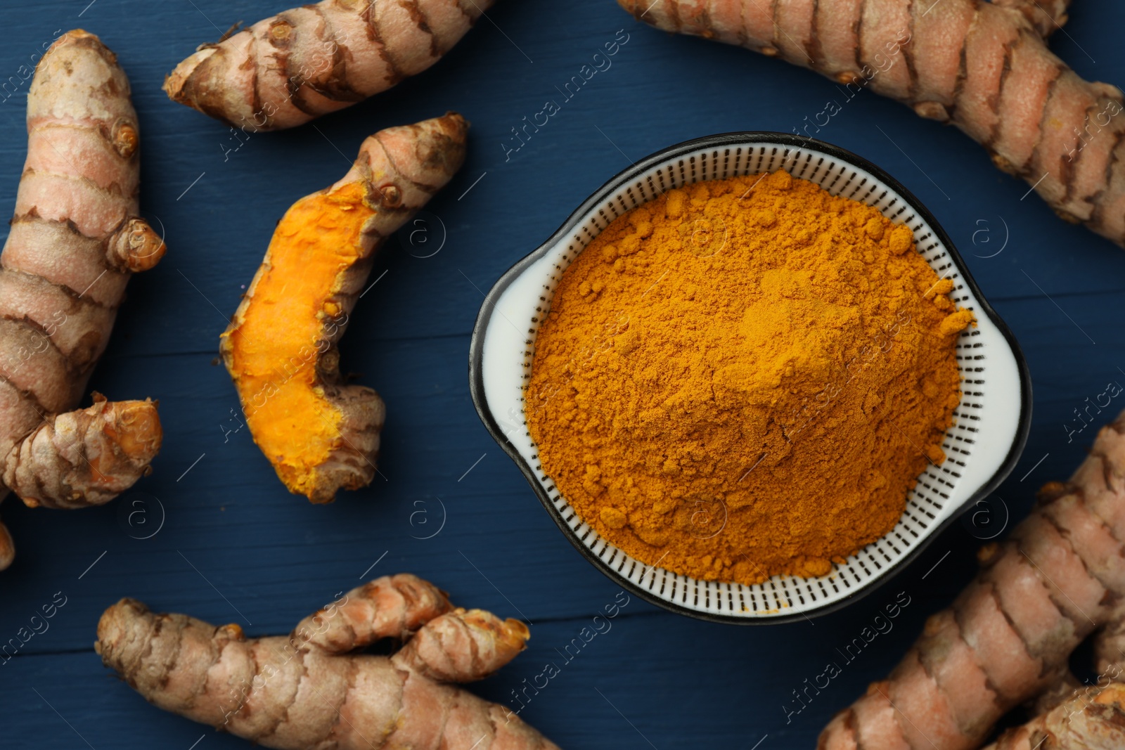 Photo of Bowl with turmeric powder and raw roots on blue wooden table, flat lay