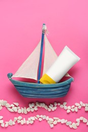 Suntan cream in toy sailboat and white marble pebbles on pink background, flat lay