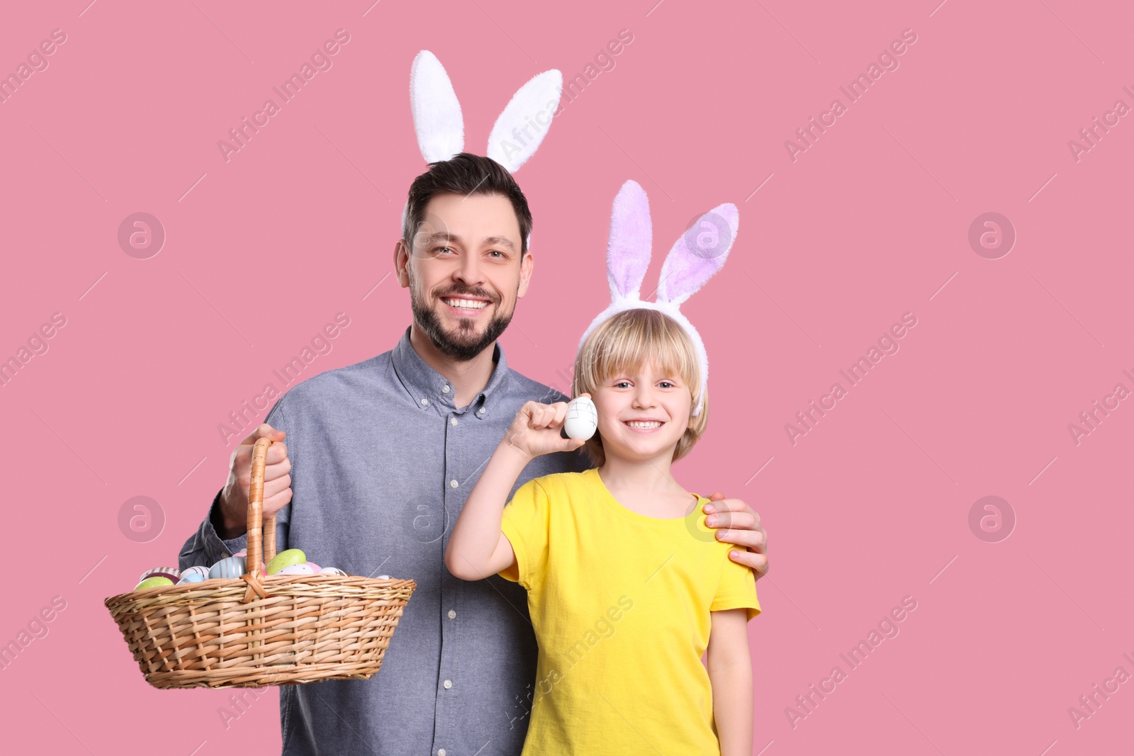 Photo of Father and son in bunny ears headbands with wicker basket of painted Easter eggs on pink background. Space for text