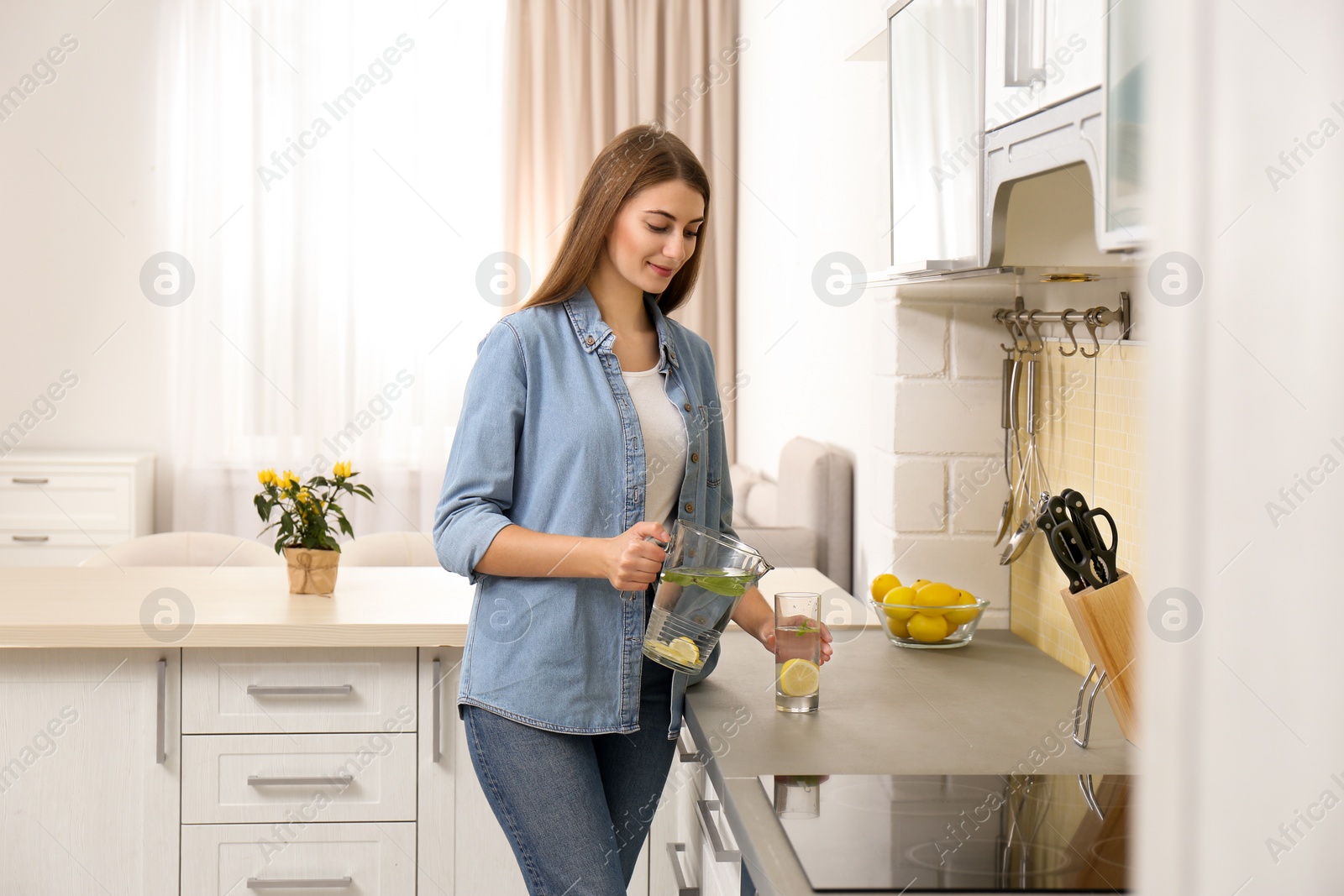 Photo of Young woman pouring lemon water into glass in kitchen