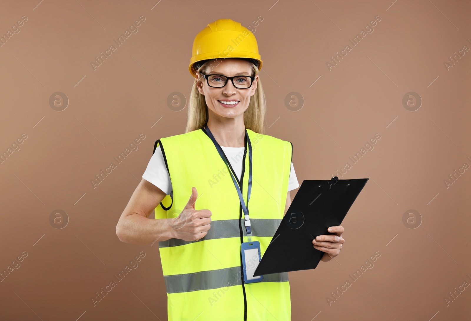 Photo of Engineer in hard hat holding clipboard and showing thumb up on brown background