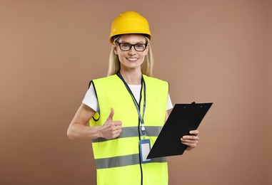 Photo of Engineer in hard hat holding clipboard and showing thumb up on brown background