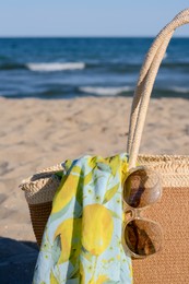 Photo of Straw bag with beach wrap and sunglasses on sandy seashore, closeup. Summer accessories