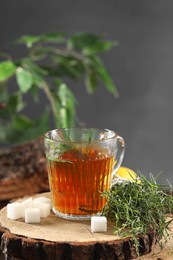 Photo of Aromatic herbal tea, fresh tarragon sprigs and sugar cubes on wooden stump, space for text