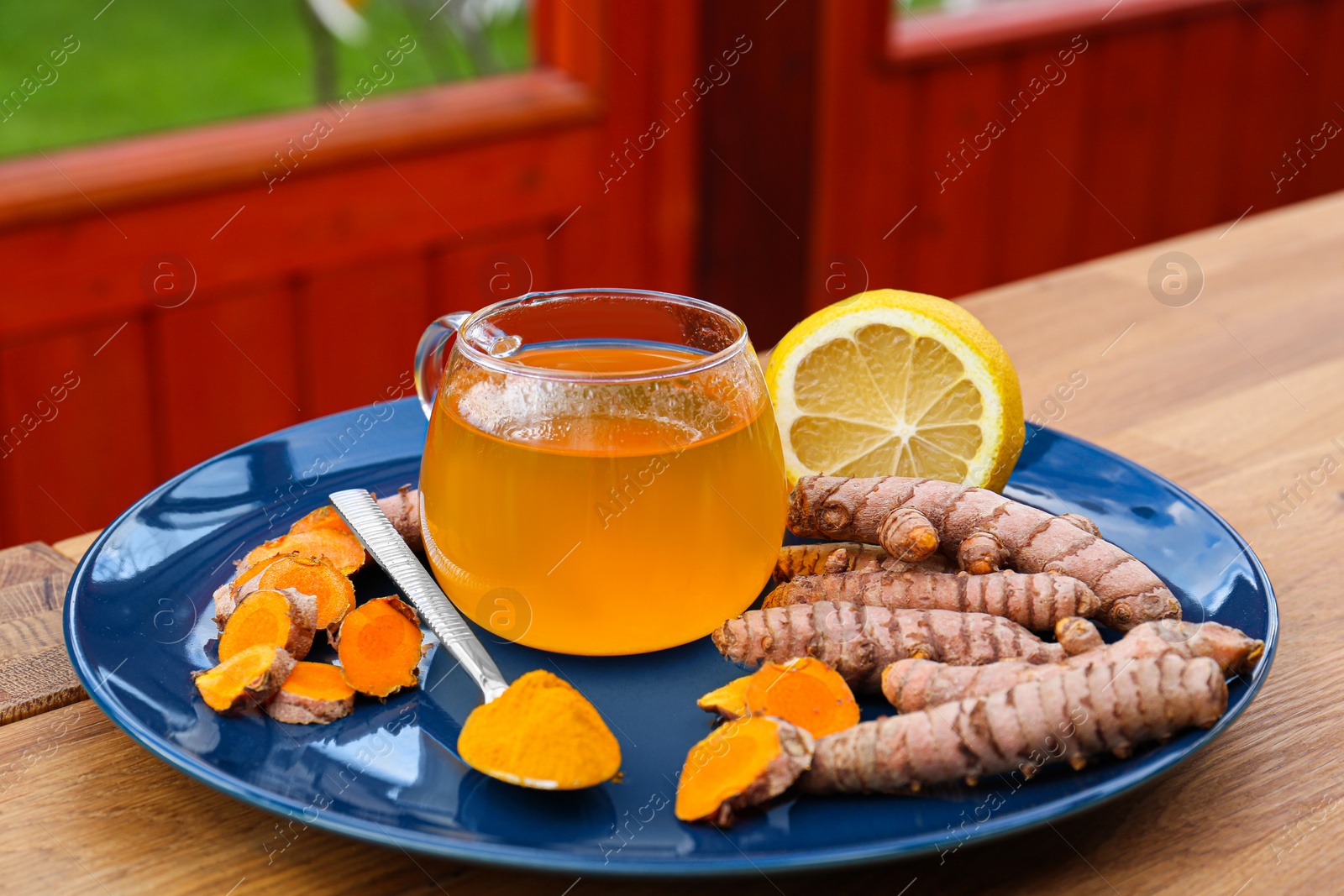 Photo of Plate with glass cup of hot tea, lemon, turmeric powder and roots on wooden table