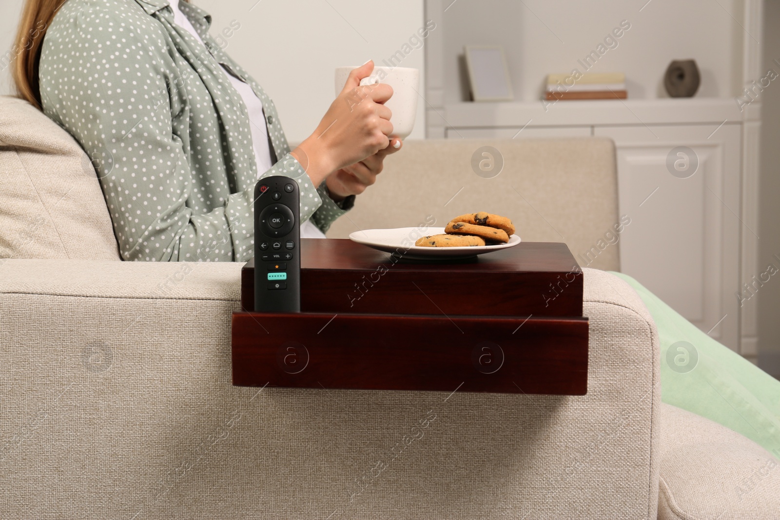 Photo of Remote control and cookies on sofa armrest wooden table. Woman holding cup of drink at home, closeup