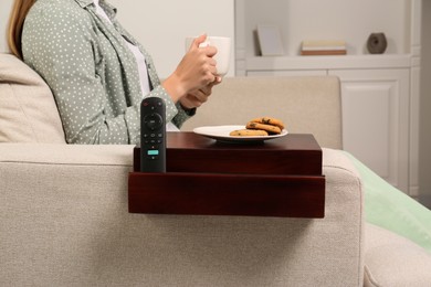 Remote control and cookies on sofa armrest wooden table. Woman holding cup of drink at home, closeup