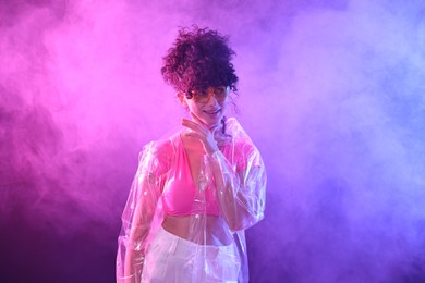 Beautiful young woman in transparent coat and sunglasses posing on color background in neon lights and smoke