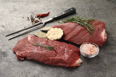 Photo of Piece of raw beef meat, spices, rosemary and fork on grey table