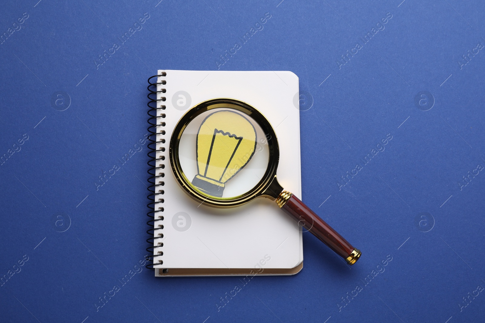 Photo of Magnifying glass over notebook with paper bulb on blue background, top view