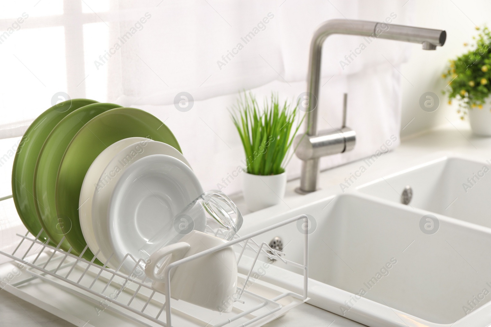 Photo of Drainer with different clean dishware, glass and cup on light table near sink indoors