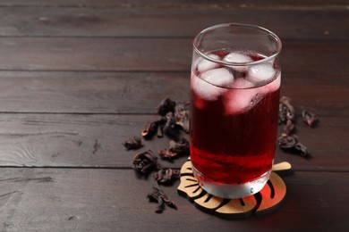 Refreshing hibiscus tea with ice cubes in glass and dry roselle petals on wooden table. Space for text
