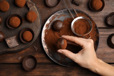 Photo of Woman covering delicious chocolate truffles with cocoa powder at wooden table, top view