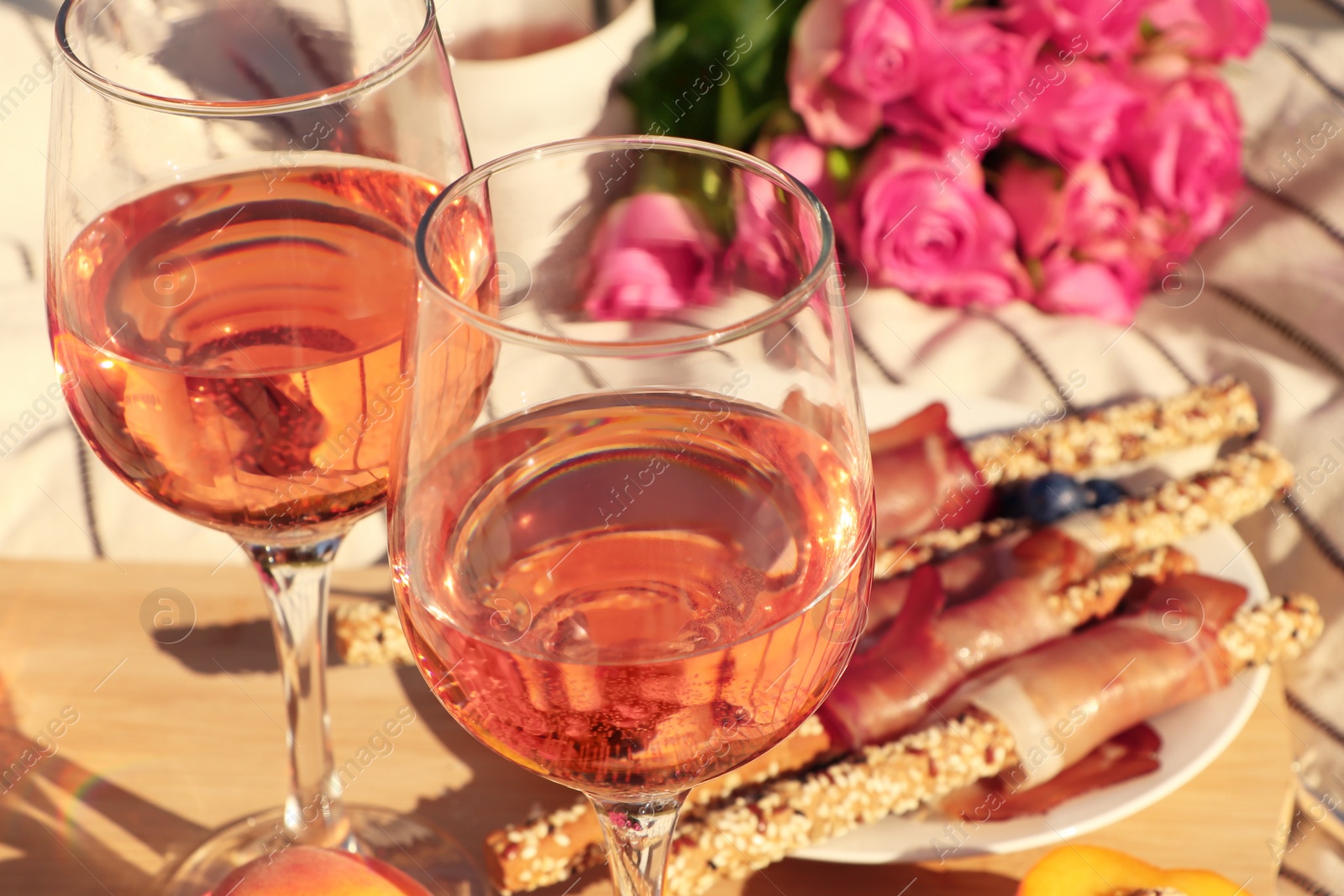 Photo of Glasses of delicious rose wine, flowers and food on white picnic blanket, closeup