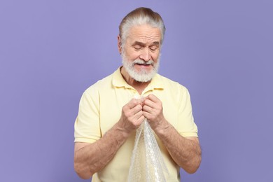 Senior man popping bubble wrap on light purple background. Stress relief