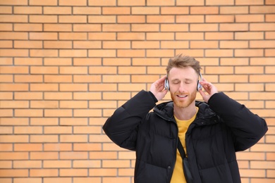 Photo of Young man listening to music with headphones against brick wall. Space for text