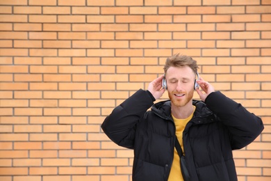 Young man listening to music with headphones against brick wall. Space for text