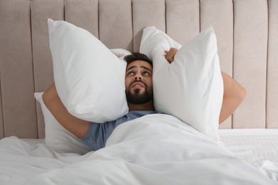 Photo of Unhappy young man covering ears with pillows in bed. Noisy neighbours