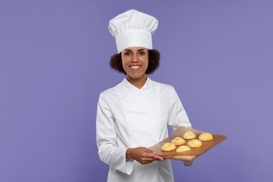 Photo of Happy female chef in uniform holding board with cookies on purple background