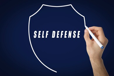 Image of Man drawing shield around words Self Defense with marker on glass board against dark blue background, closeup