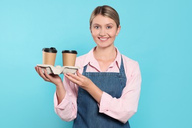 Photo of Beautiful young woman in denim apron with cups of coffee on light blue background