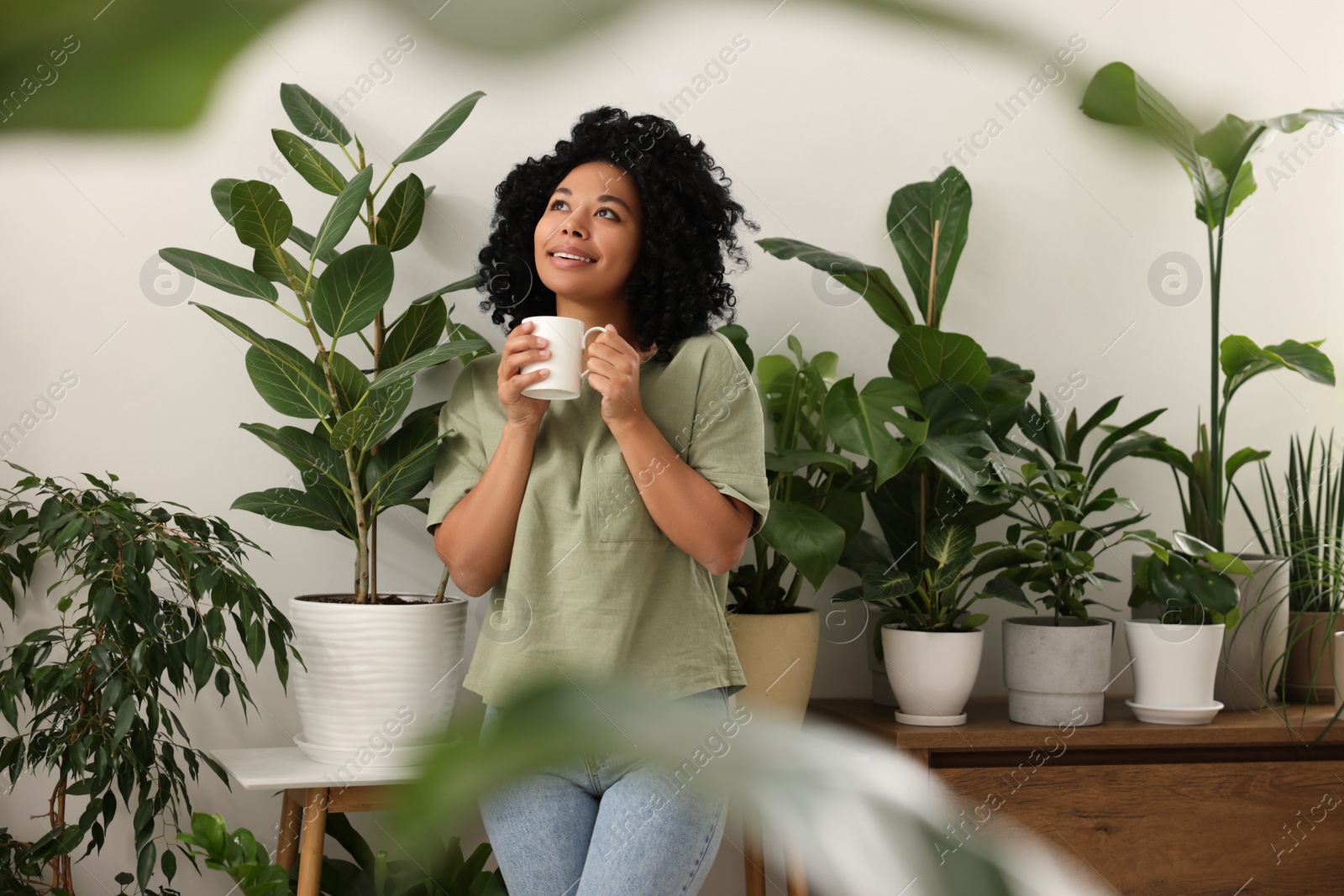 Photo of Relaxing atmosphere. Happy woman with cup of hot drink near beautiful houseplants in room