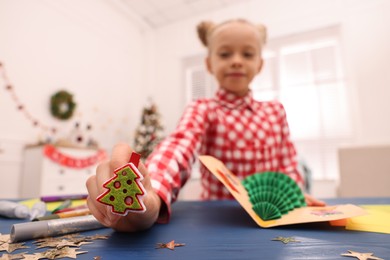 Photo of Cute little child with beautiful Christmas card at table in room, focus on decorative clothespin