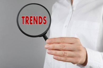 Image of Searching new and popular trends. Woman holding magnifying glass over word on grey background, closeup