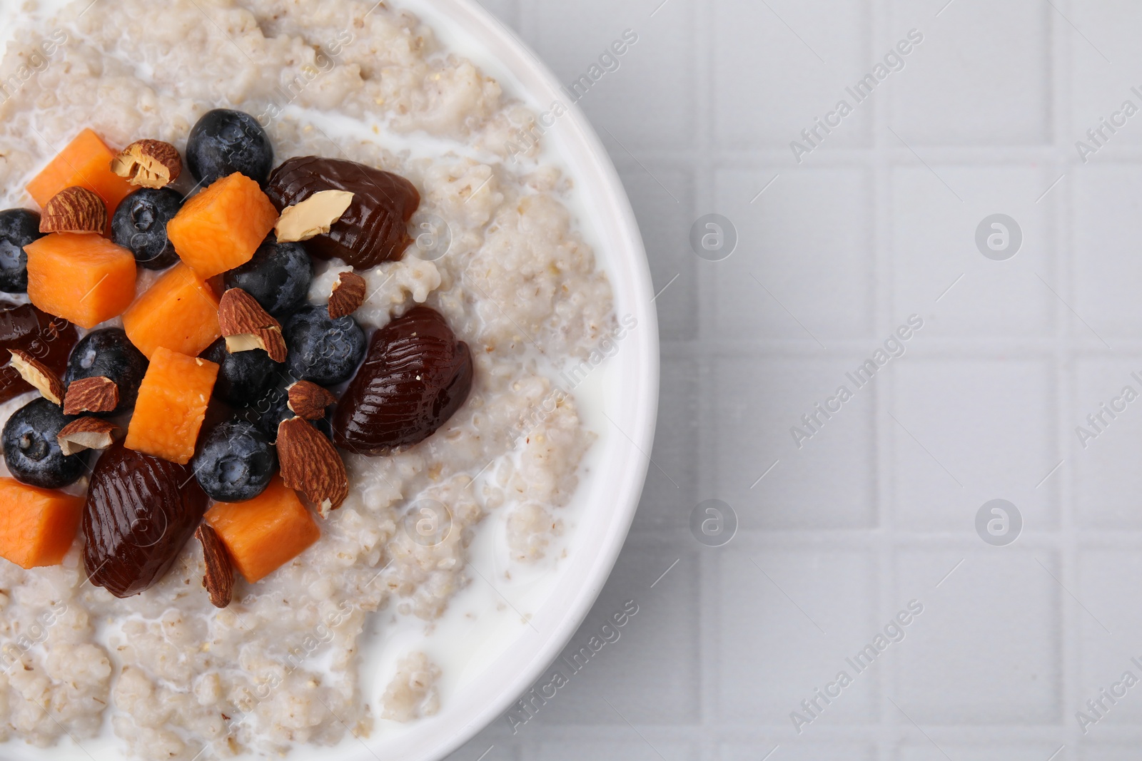 Photo of Delicious barley porridge with blueberries, pumpkin, dates and almonds in bowl on white tiled table, top view. Space for text