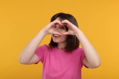 Photo of Happy woman looking through folded in shape of heart hands on orange background