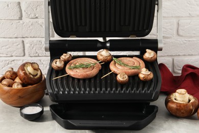 Photo of Electric grill with homemade sausages, rosemary and mushrooms on marble table