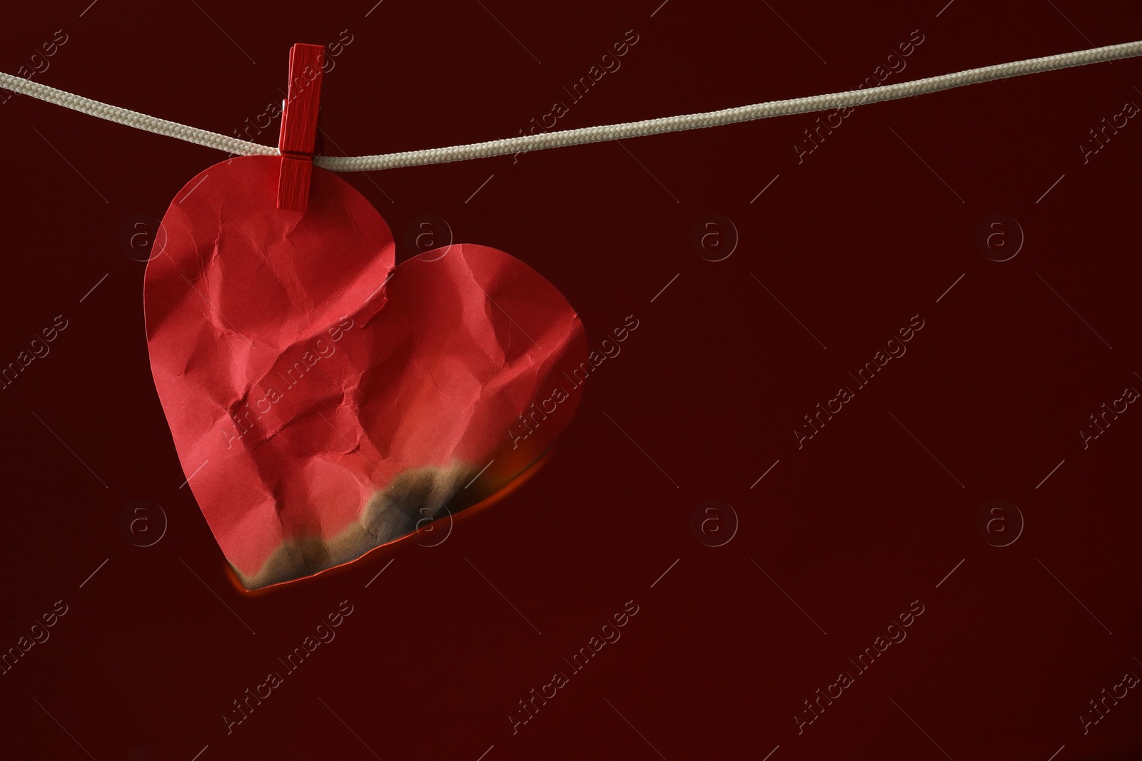 Photo of Crumpled red paper heart burning on rope against burgundy background, space for text. Broken heart