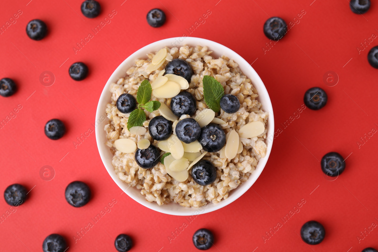 Photo of Tasty oatmeal with blueberries, mint and almond petals in bowl surrounded by fresh berries on red background, flat lay