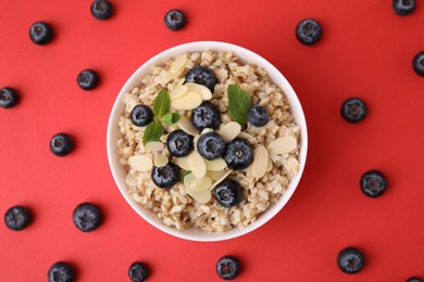 Photo of Tasty oatmeal with blueberries, mint and almond petals in bowl surrounded by fresh berries on red background, flat lay