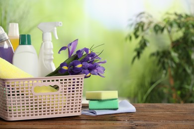 Spring cleaning. Plastic basket with detergents, supplies and beautiful flowers on wooden table outdoors, space for text