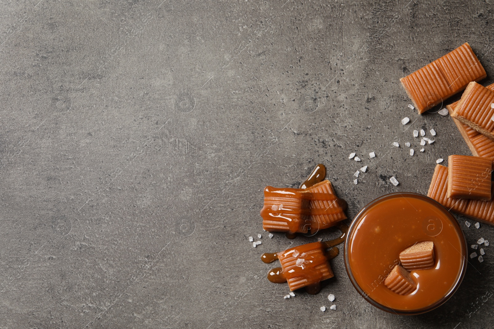 Photo of Tasty salted caramel with candies in glass bowl on grey table, flat lay. Space for text