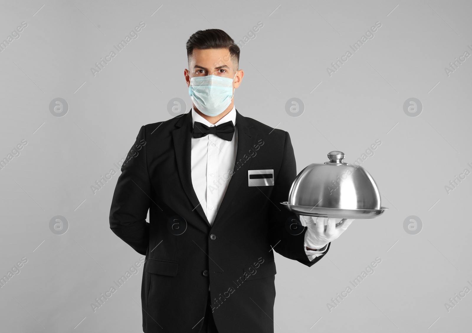 Photo of Waiter in medical face mask holding tray with lid on light grey background