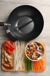 Photo of Black wok, chopsticks and board with products on light wooden table, top view