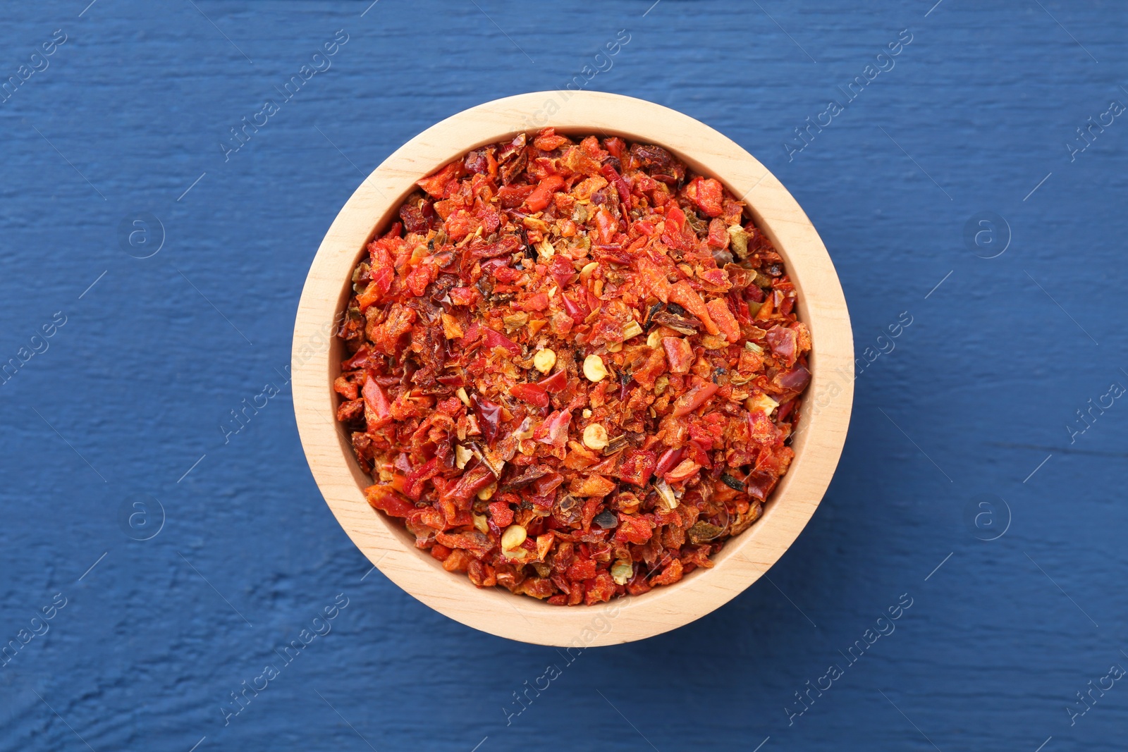 Photo of Chili pepper flakes in bowl on blue wooden table, top view