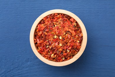 Photo of Chili pepper flakes in bowl on blue wooden table, top view