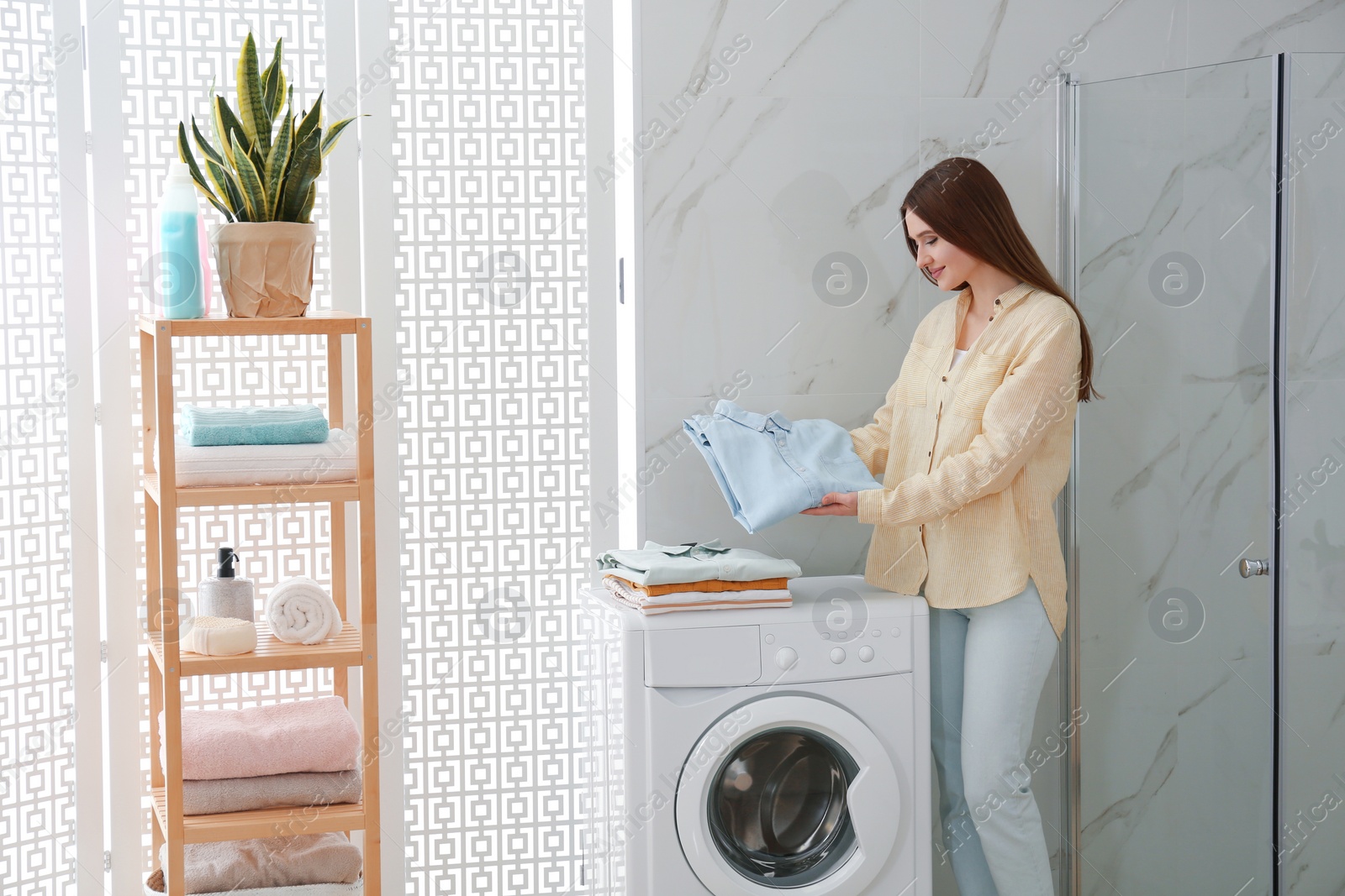 Photo of Happy woman with clean clothes near washing machine in bathroom. Laundry day