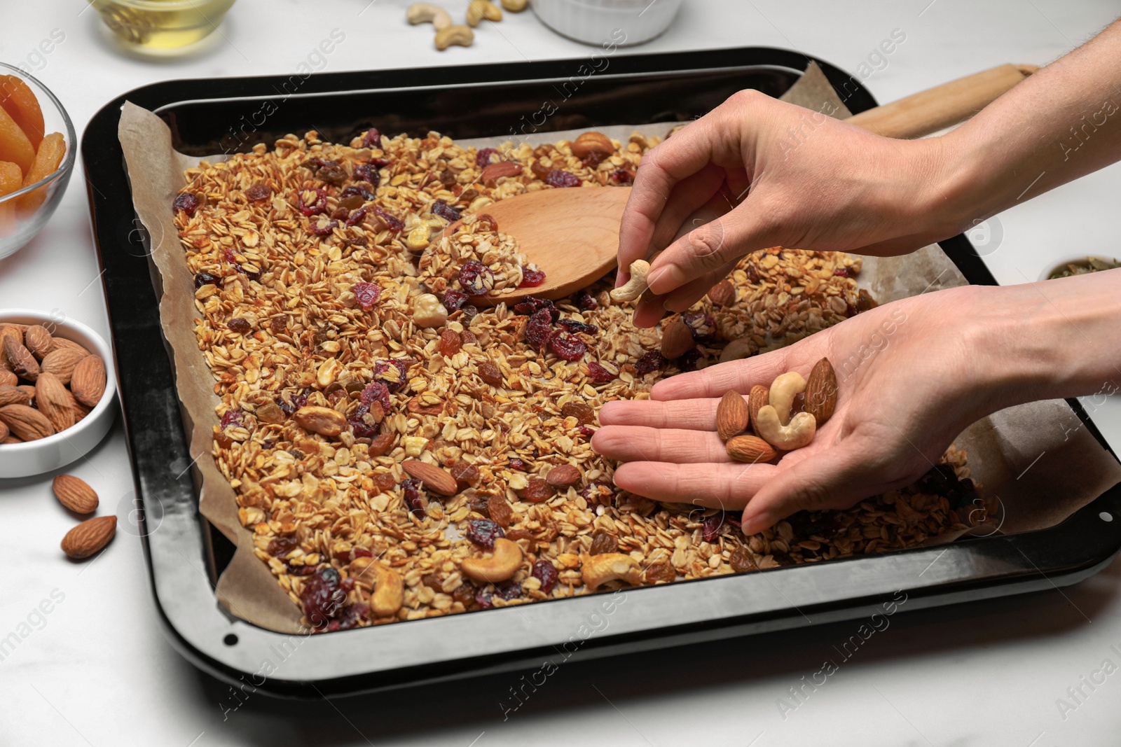 Photo of Woman putting nuts into tray with tasty granola at white marble table, closeup